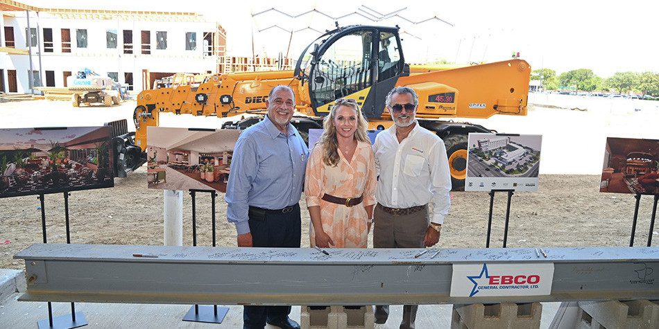 Two men and a woman standing behind the final beam for Caravan Court Hotel, in front of a bulldozer
